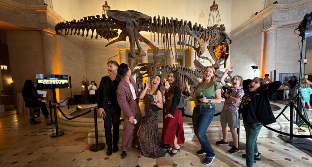 Players on a scavenger hunt at the Natural History Museum pose with dinosaurs.