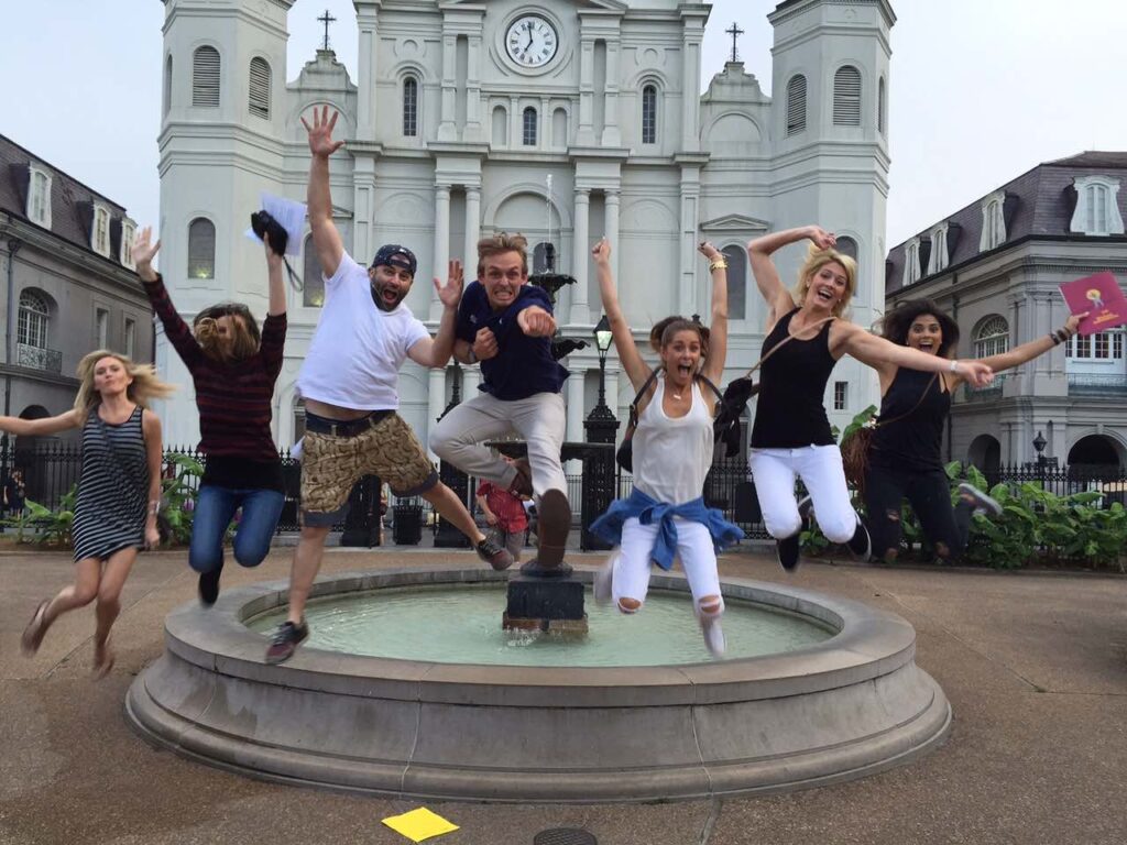 7 friends on a scavenger hunt jump in front of fountain and white building with a clock 